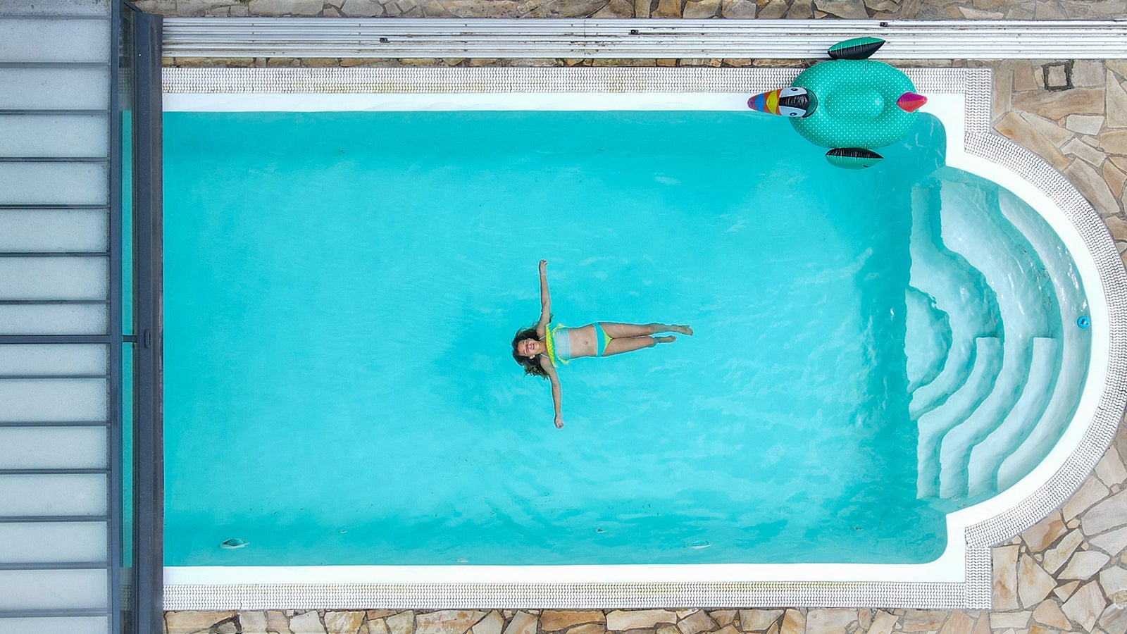 ILLUSTRATION - 06.07.2022, Brandenburg, Mixdorf: ARCHIV - Ein Kind schwimmt in einem Pool, w&auml;hrend ein anderes M&auml;dchen den Rasen mit einem Wasserschlauch beregnet (Luftaufnahme mit einer Drohne, gestellte Aufnahme). Wenn es hei&szlig; ist, einfach in den Garten gehen und ins kalte Wasser springen! Pools liegen im Trend. Das st&ouml;&szlig;t nicht &uuml;berall auf Begeisterung. (zu dpa "Ausgeplanscht? - Private Pools wegen Wassermangels in der Kritik") Foto: Patrick Pleul/dpa +++ dpa-Bildfunk +++
