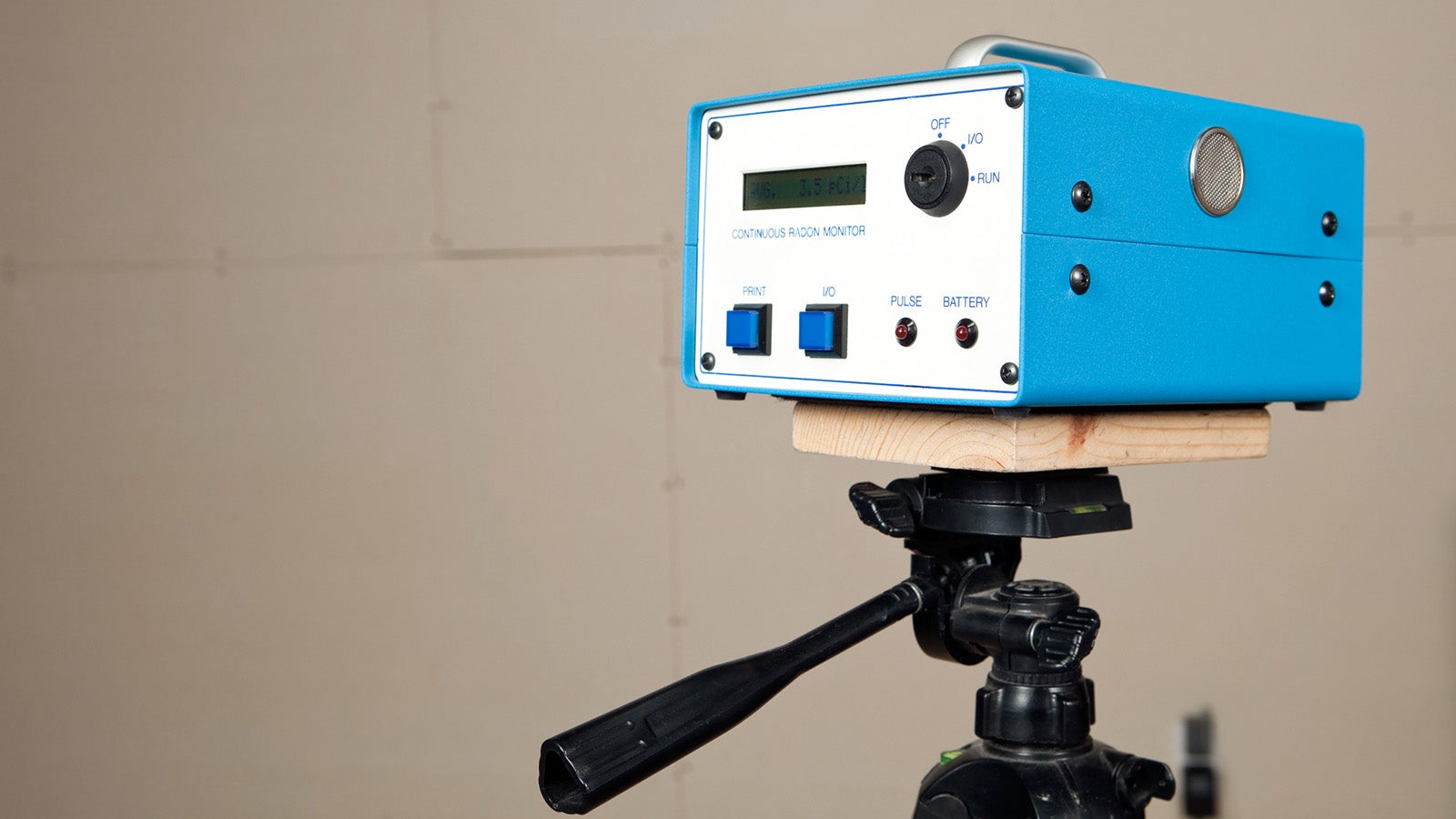 "A continuous radon monitor is mounted to a tripod in an unfinished house basement. A house is commonly tested for radon with a single use test kit. If initial readings are high or remediation work has begun a system like this would be used to determine a more accurate measurement over time. An added benefit of this unit is real time measurement. The display screen reads Avg. 3.5 pCi/l  (picocuries per liter), the curie is a unit of radioactivity. The background is unfinished drywall with seams and screws."