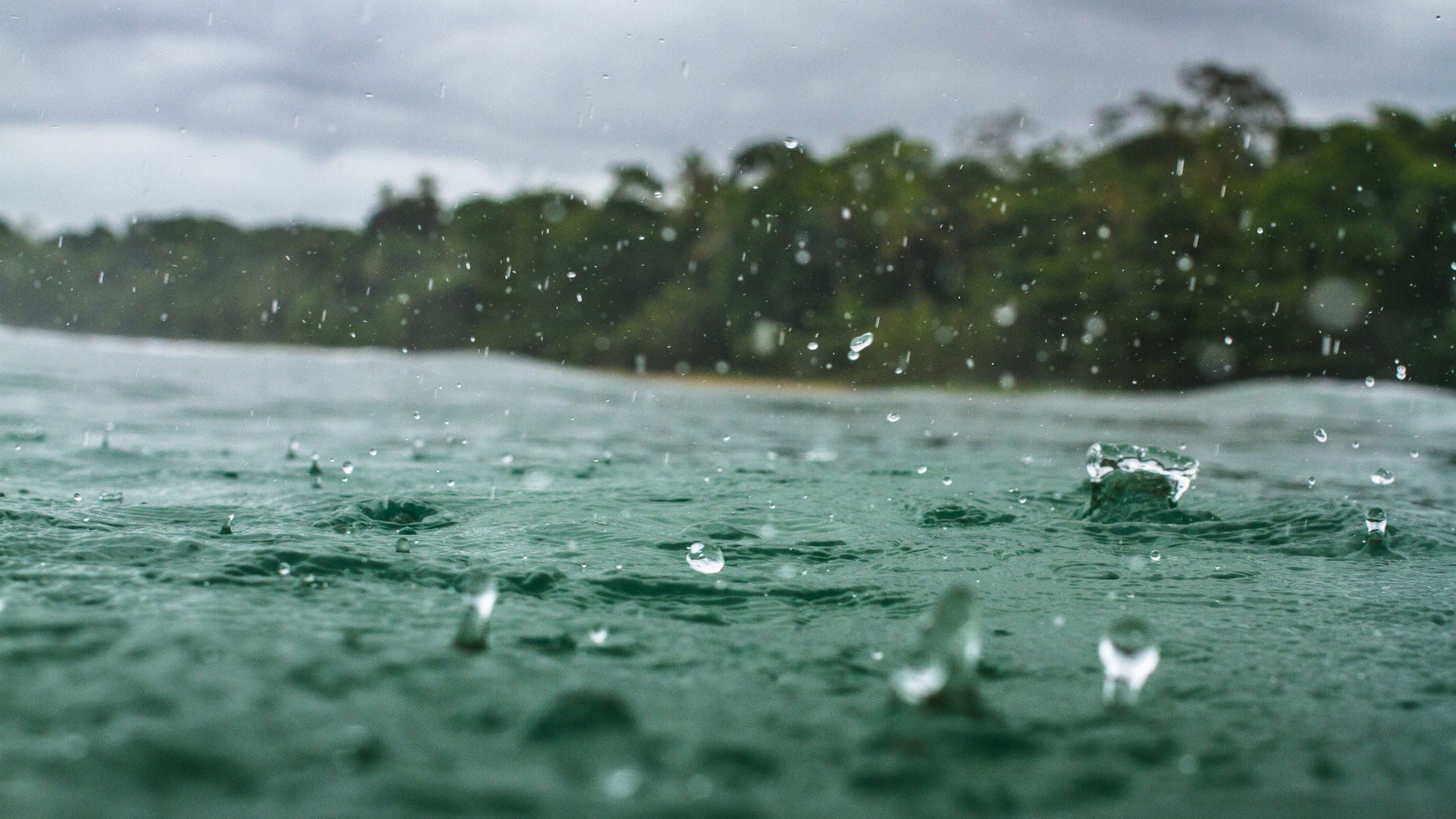 Rain falls on the water of a beautiful beach in the Caribbean, a stretch of a jungle laden coastline can be seen in the background.