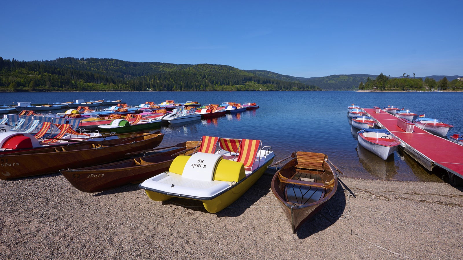 Mehrere Tretboote und Ruderboote liegen am Ufer eines Sees neben einem Steg