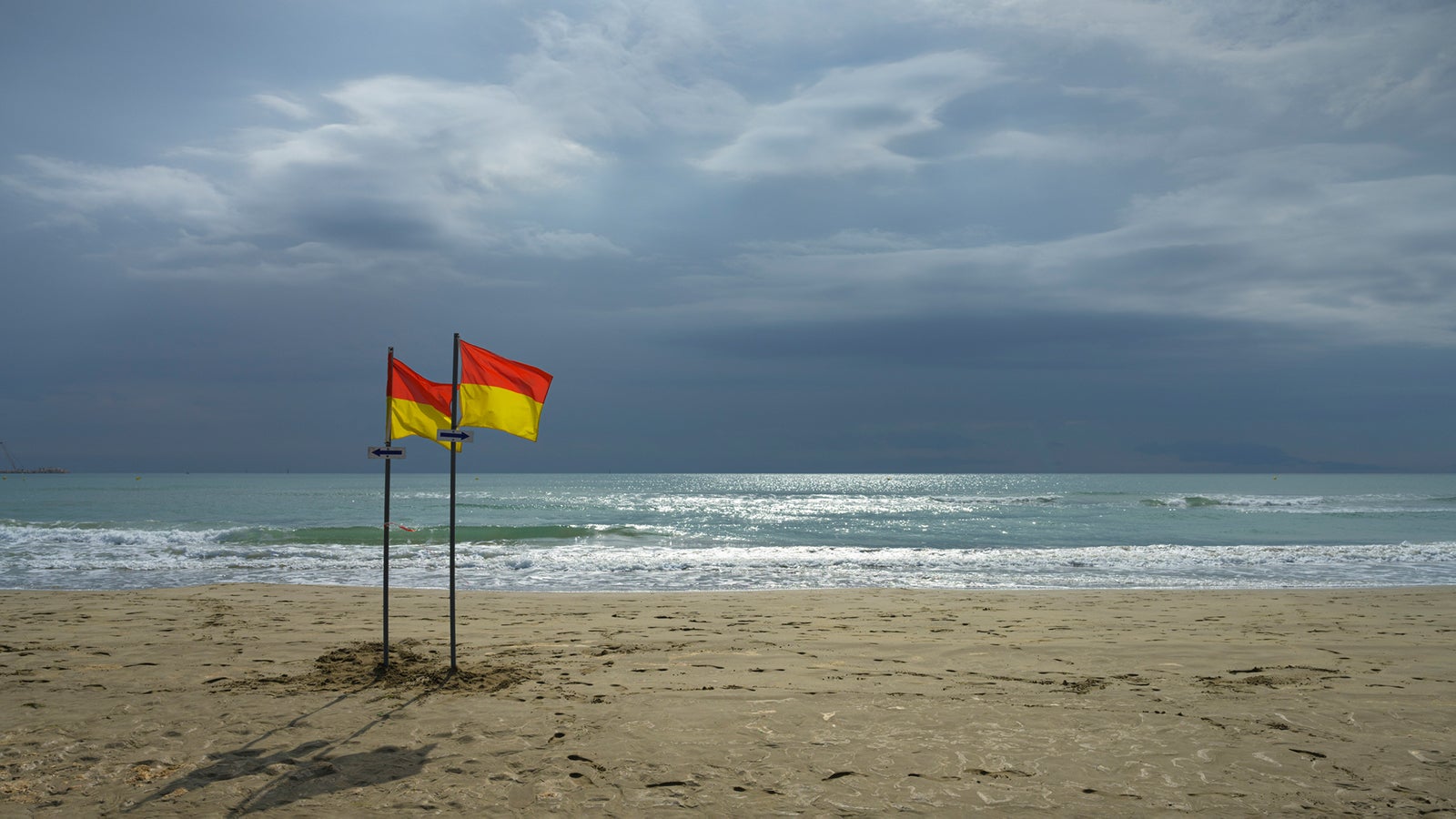 Zwei rot-gelbe Flaggen auf Stangen stehen am menschenleeren Sandstrand vor ruhigem Meer und bew&ouml;lktem Himmel in Port-La-Nouvelle, Frankreich