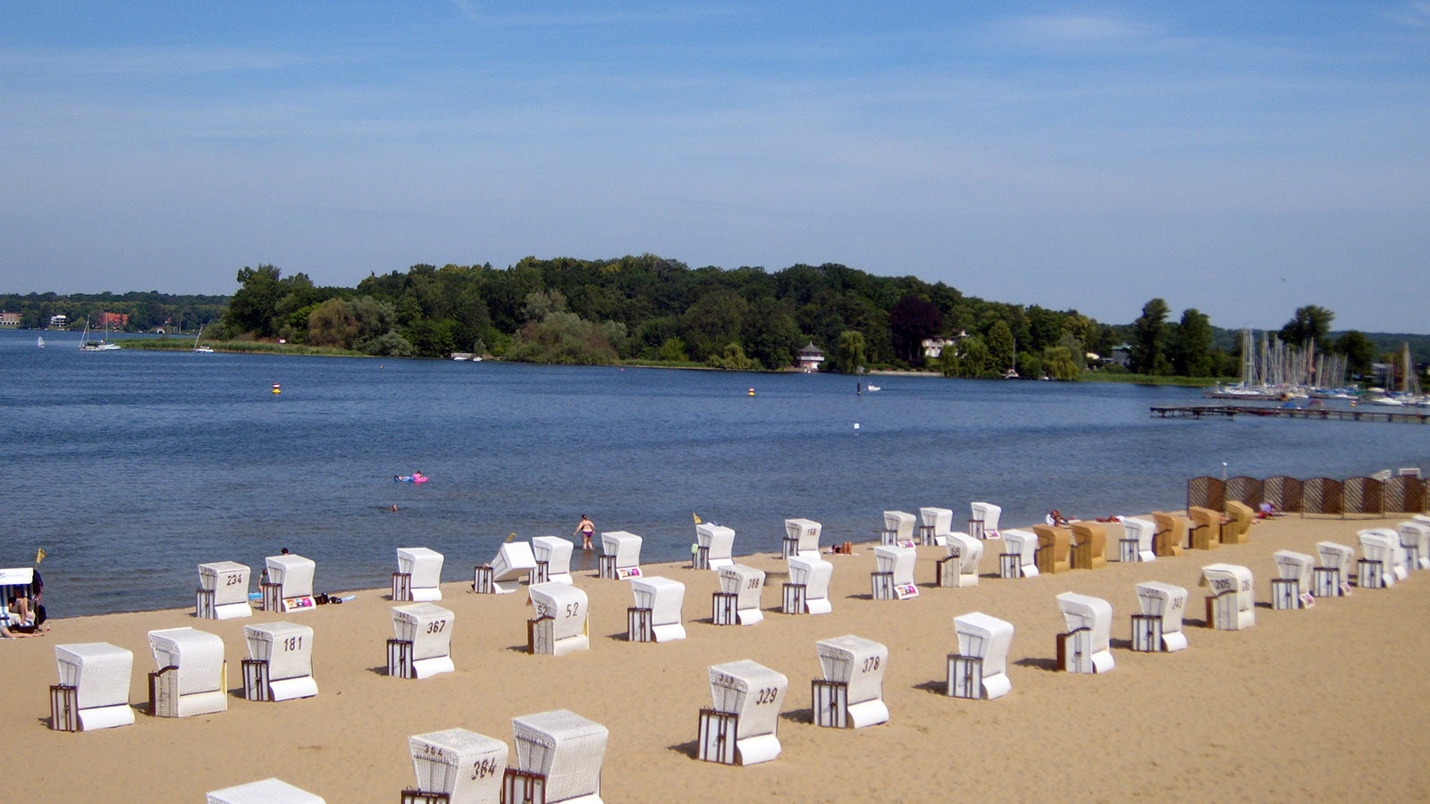 Strand mit mehreren Strandk&ouml;rben vor einem ruhigen See und bewaldetem Ufer im Hintergrund.