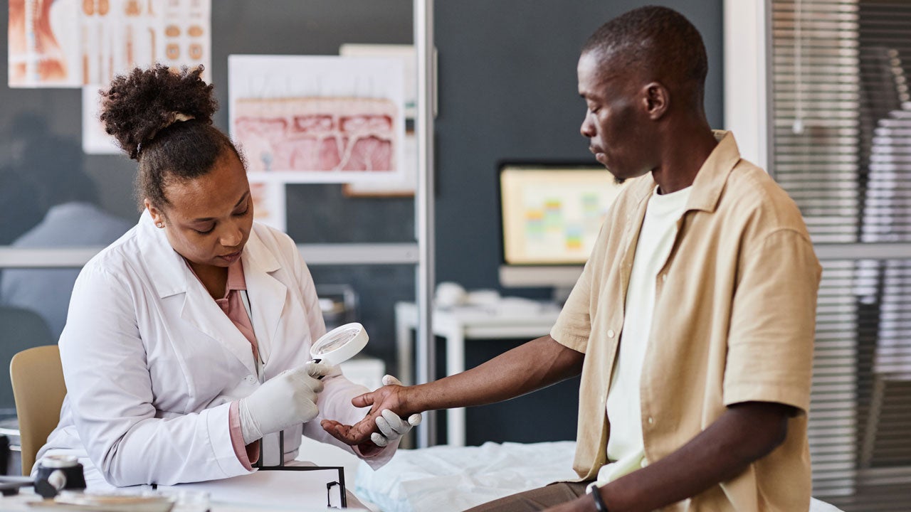 A dermatologist inspects a male patient's arm with a magnifying glass 