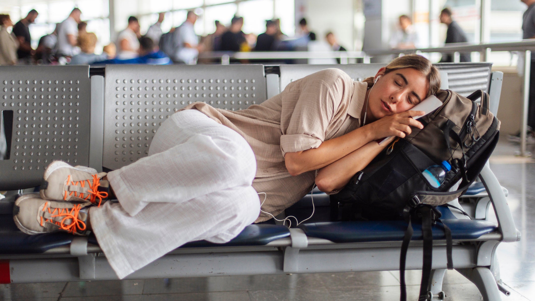 A young woman naps on an airport bench, ears in headphones, clutching a phone, with a backpack beside her; The busy terminal hums in the background as travelers move past