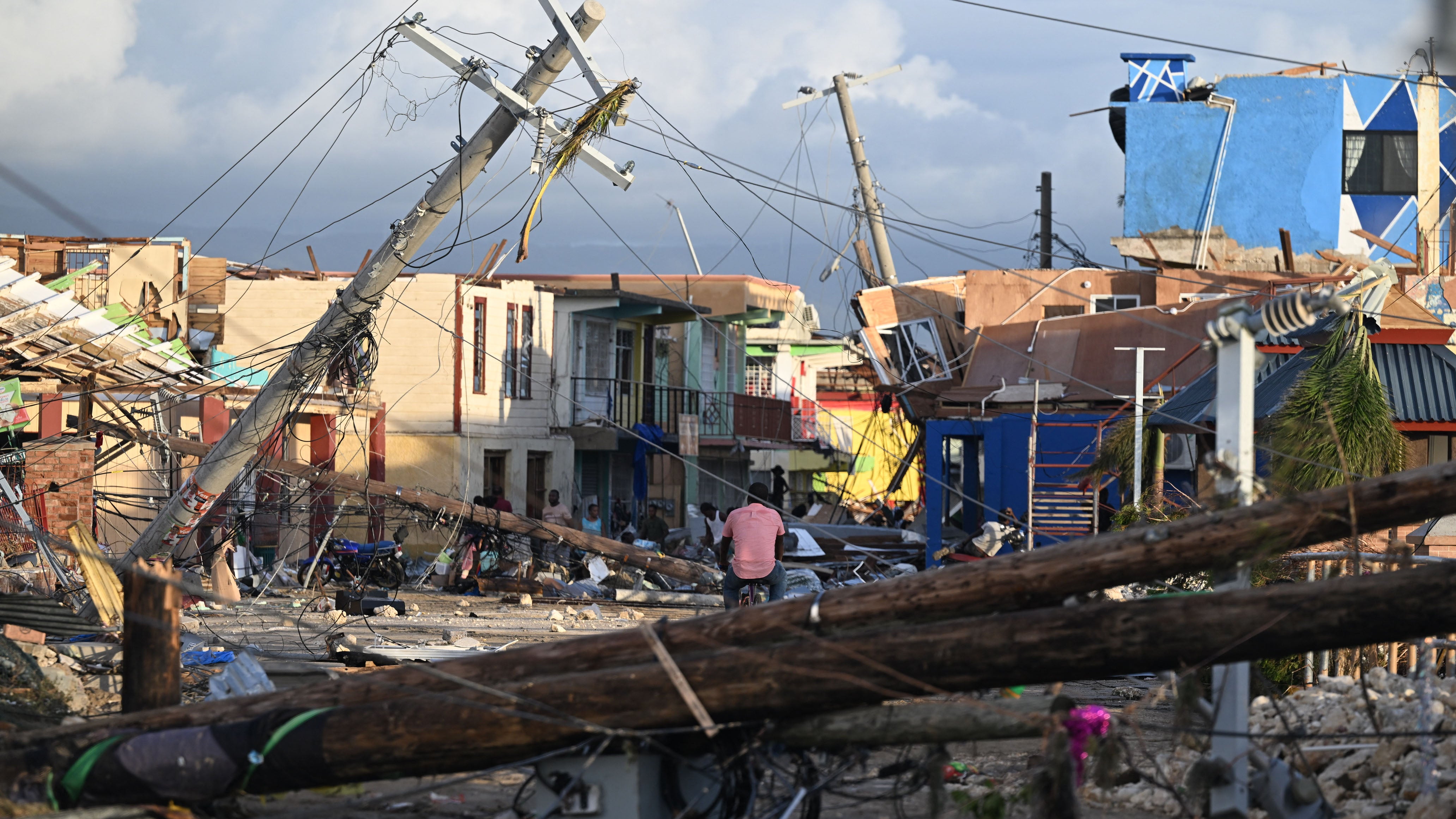 TOPSHOT - Electrical poles are down as a man bikes through the destroyed neighborood of North Street following the passage of Hurricane Melissa, in Black River, Jamaica on October 29, 2025. Hurricane Melissa bore down on the Bahamas October 29 after cutting a path of destruction through the Caribbean, leaving 30 people dead or missing in Haiti and parts of Jamaica and Cuba in ruins. Somewhat weakened but still threatening, Melissa will bring damaging winds and flooding rains to the Bahamas Wednesday before moving on to Bermuda late Thursday, according to the US National Hurricane Center (NHC). (Photo by Ricardo MAKYN / AFP) (Photo by RICARDO MAKYN/AFP via Getty Images)          