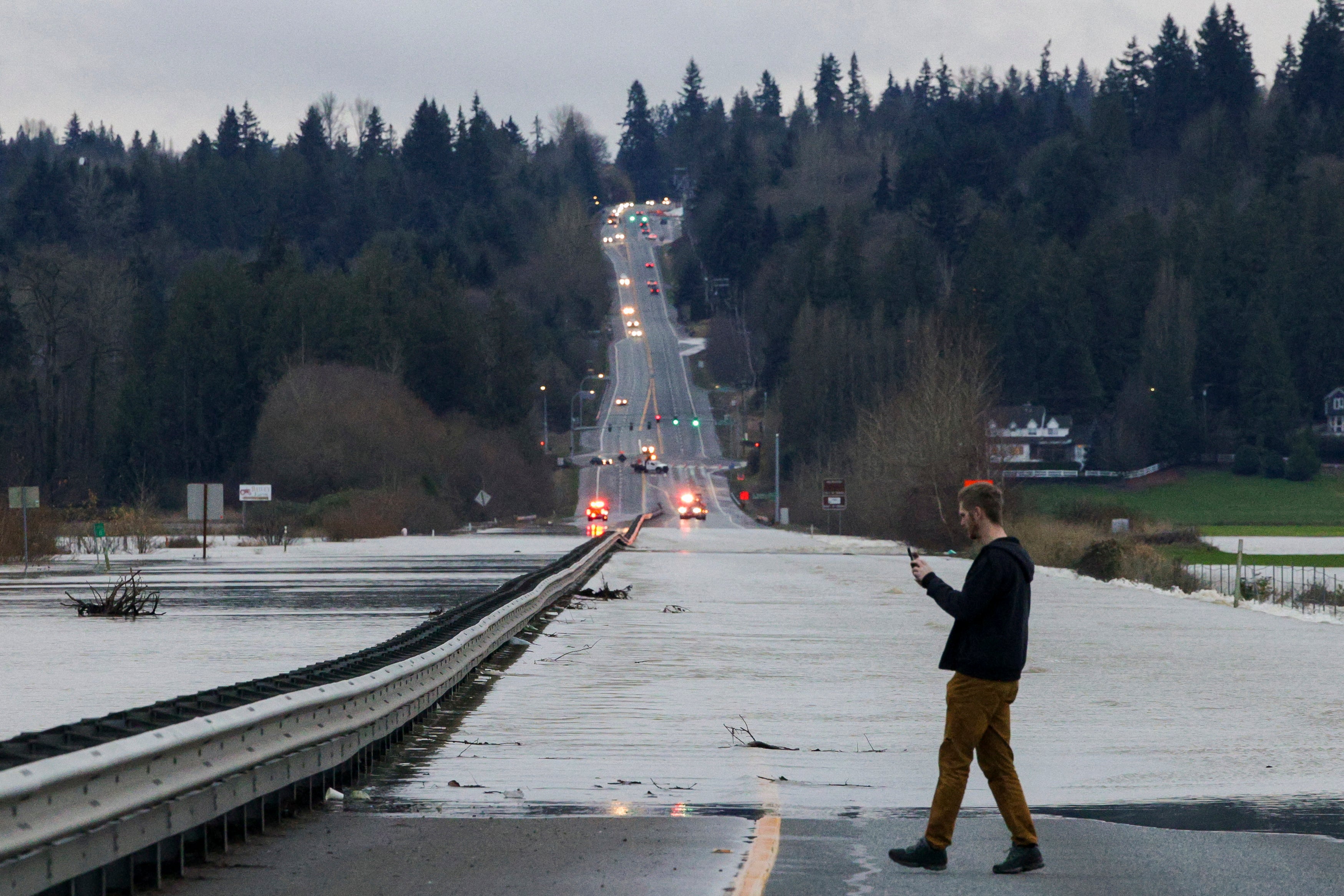 A person stops to capture cellphone images as flood waters from the Snohomish River cover a portion of State Route 9 in Snohomish, Washington, on December 11, 2025. Tens of thousands of people were under evacuation orders Thursday in western North America, after days of heavy rain forced rivers to burst their banks. Storms have battered Washington state in the US and British Columbia over the Canadian border for several days, with rivers continuing to rise. (Photo by Jason Redmond / AFP via Getty Images)