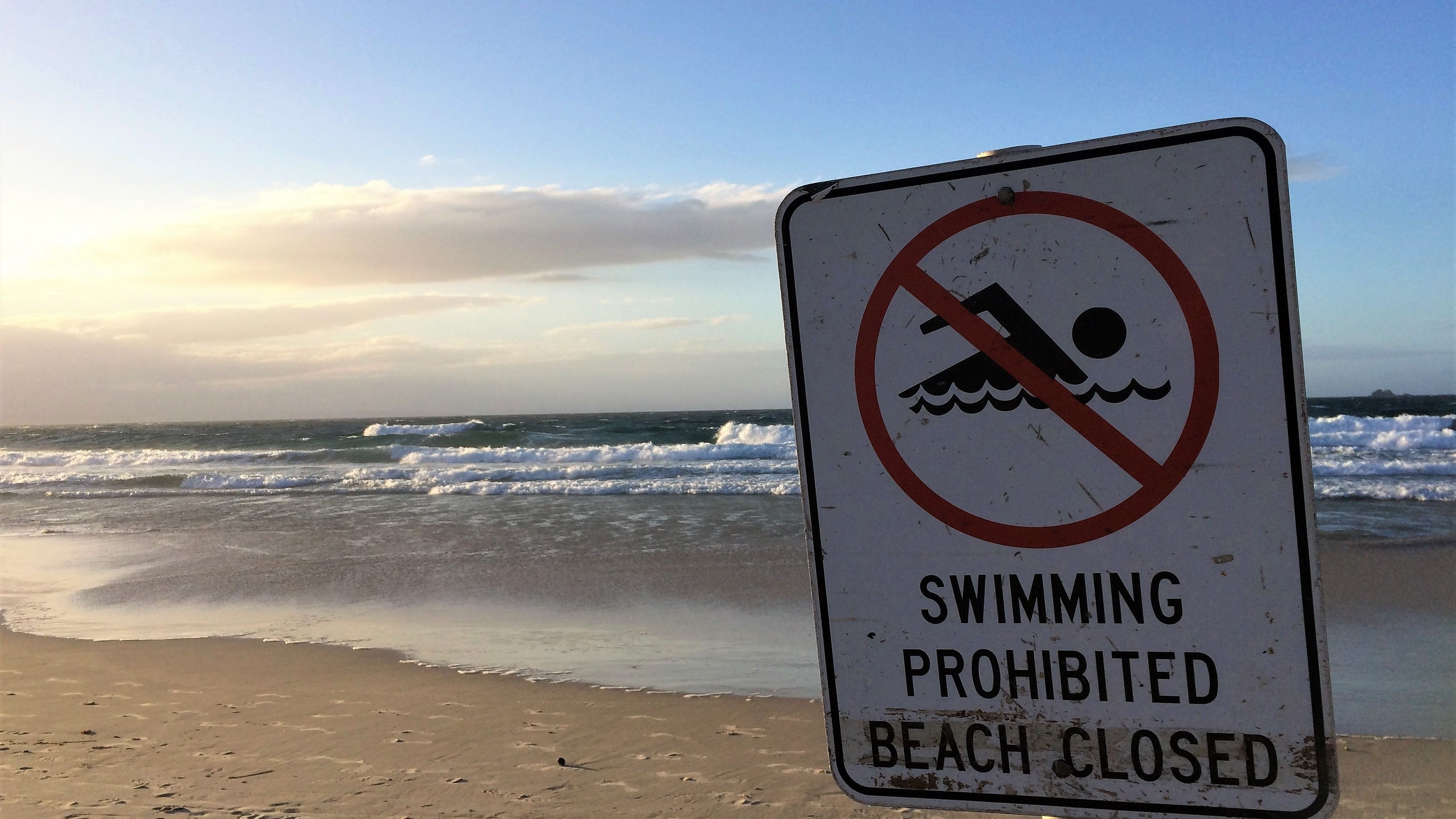 A sign on a pretty beach shows a swimmer in a circle with a red line through it and the words: 'swimming prohibited, beach closed'. The sun is setting in the distance and the day is about to end. The photo was taken in Bryon Bay, Australia.