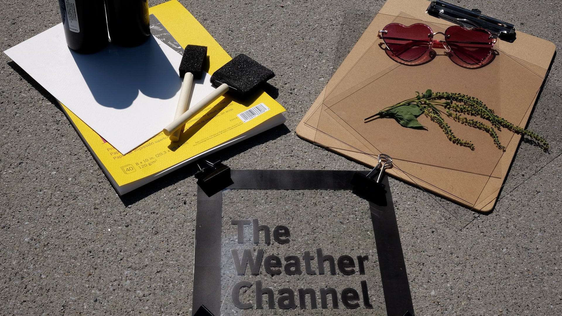 Art supplies, heart-shaped pink sunglasses, dried plant, and a clear "The Weather Channel" sign on pavement