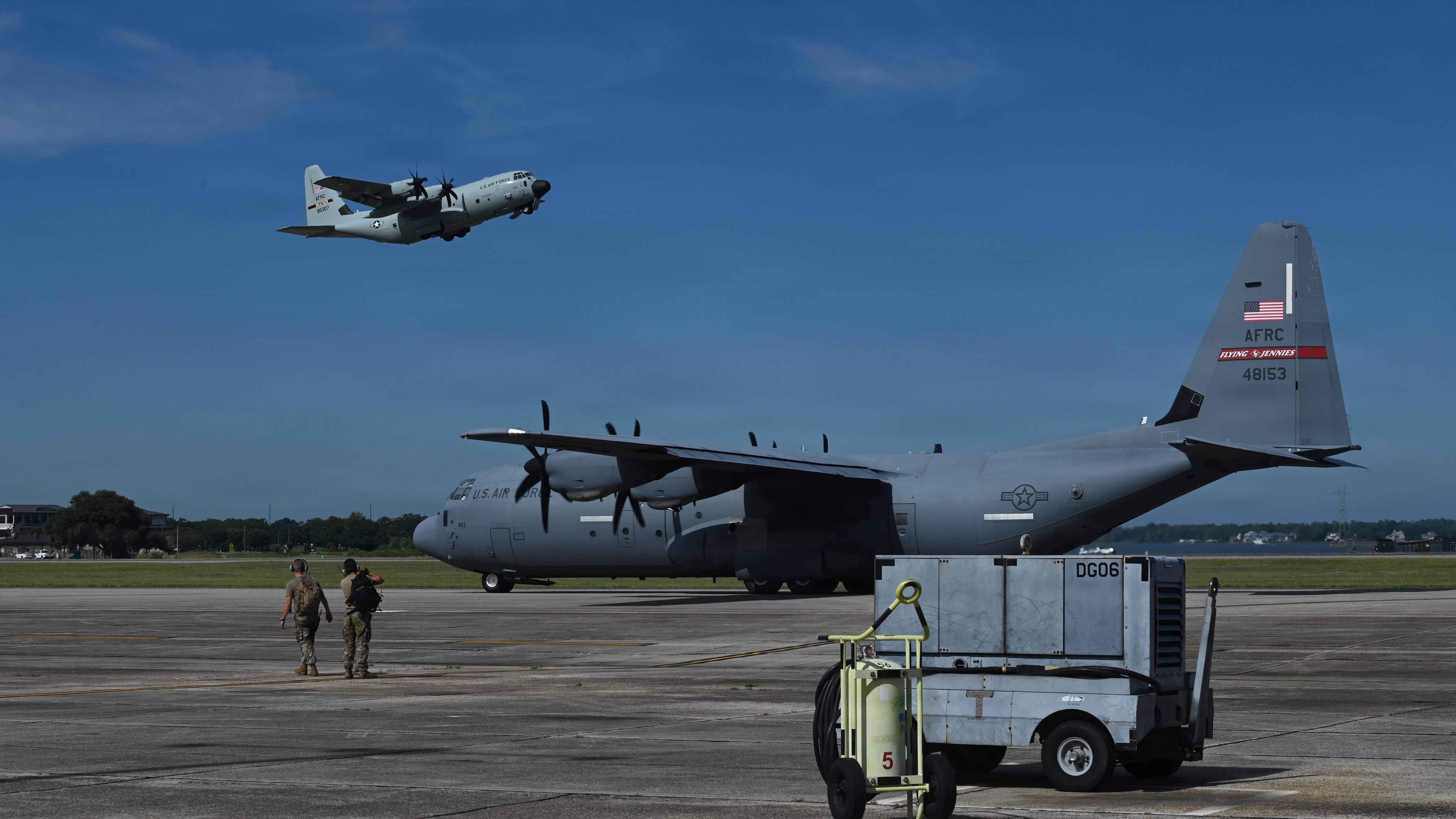 Two airmen watch a U.S. Air Force C-130 aircraft take off while another sits parked on the tarmac.