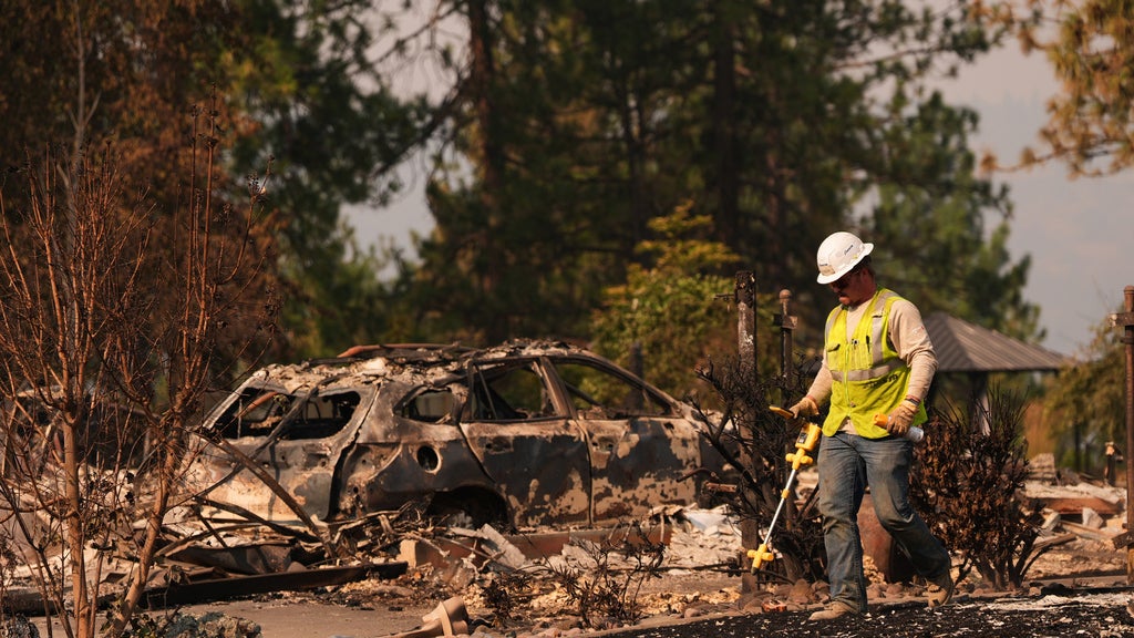 Worker in hard hat and safety vest inspects debris near a burned-out car after a wildfire.