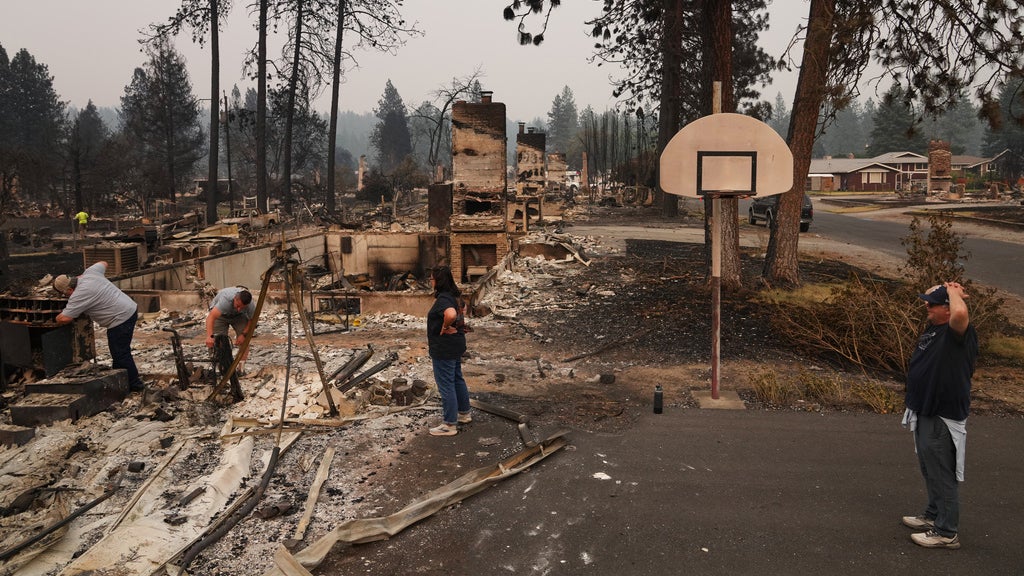 People stand amid burned-out homes and debris after a wildfire, with a still-standing basketball hoop nearby.