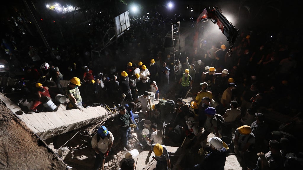Nighttime rescue workers in helmets search through rubble at a collapsed building disaster site.