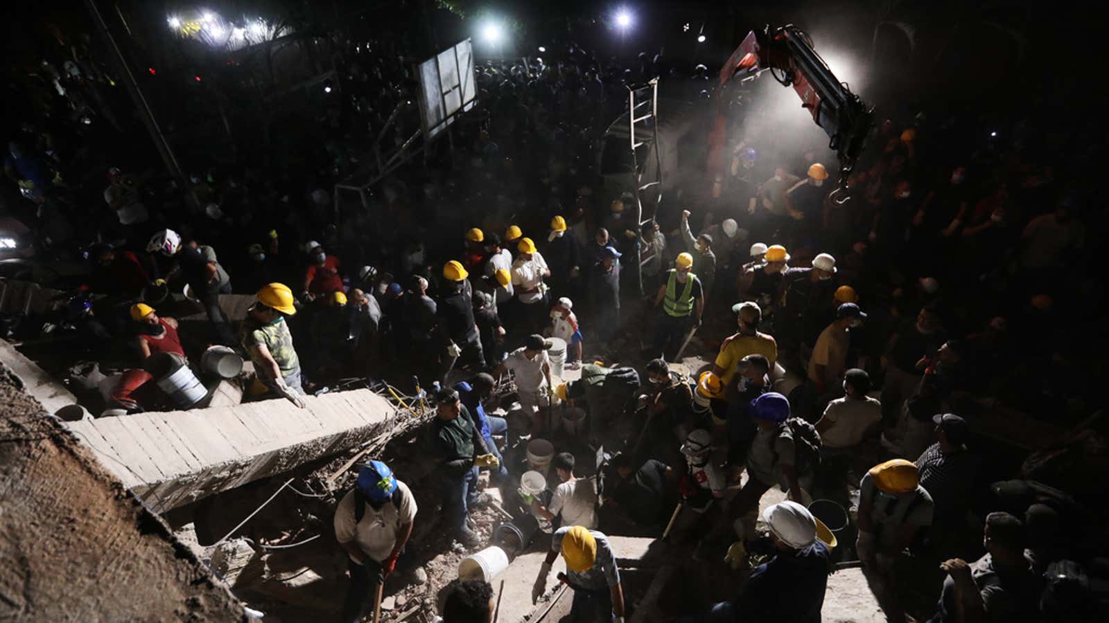 Nighttime rescue workers in helmets search through rubble at a collapsed building disaster site.