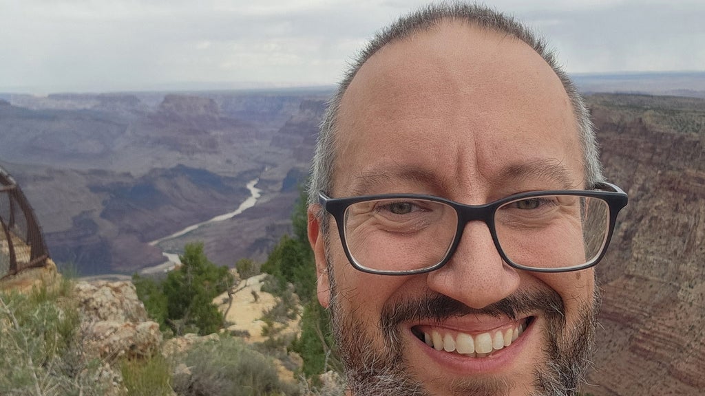 Smiling man wearing glasses takes a selfie overlooking a canyon with a winding river below.