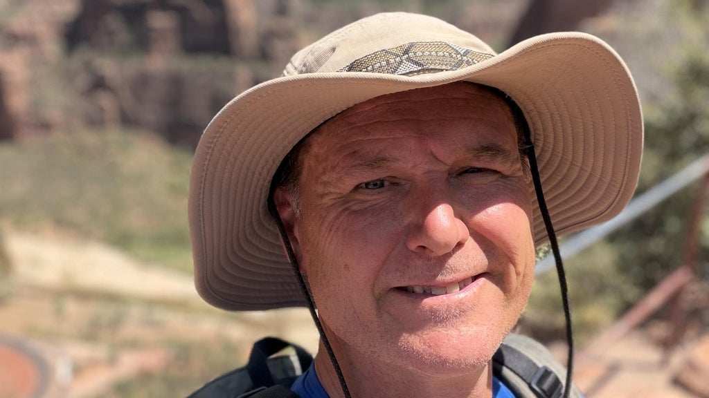 Smiling man wearing a wide-brimmed tan hat with chin strap, outdoors with blurred rocky landscape background.