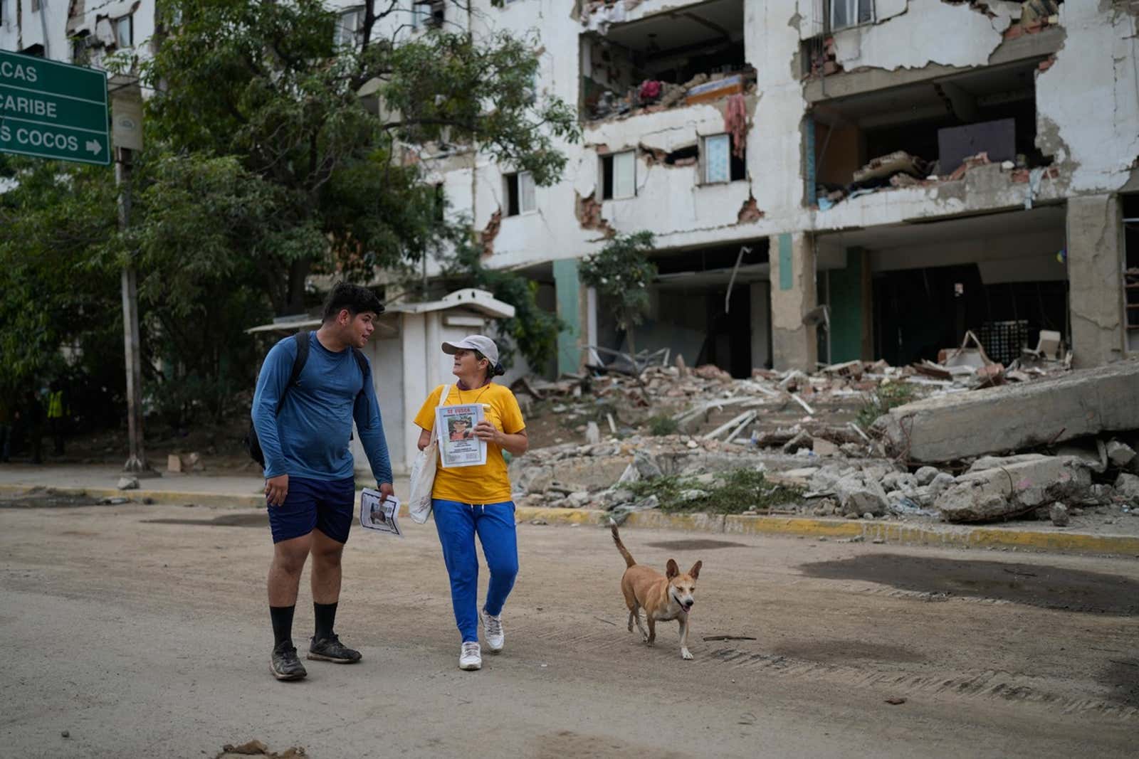 people and dog stand in front of destroyed building