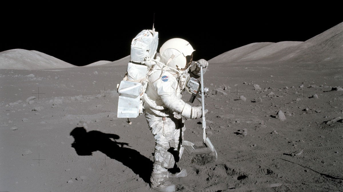 Astronaut in a white spacesuit on the lunar surface using a tool to collect lunar samples, with rocky hills in the background