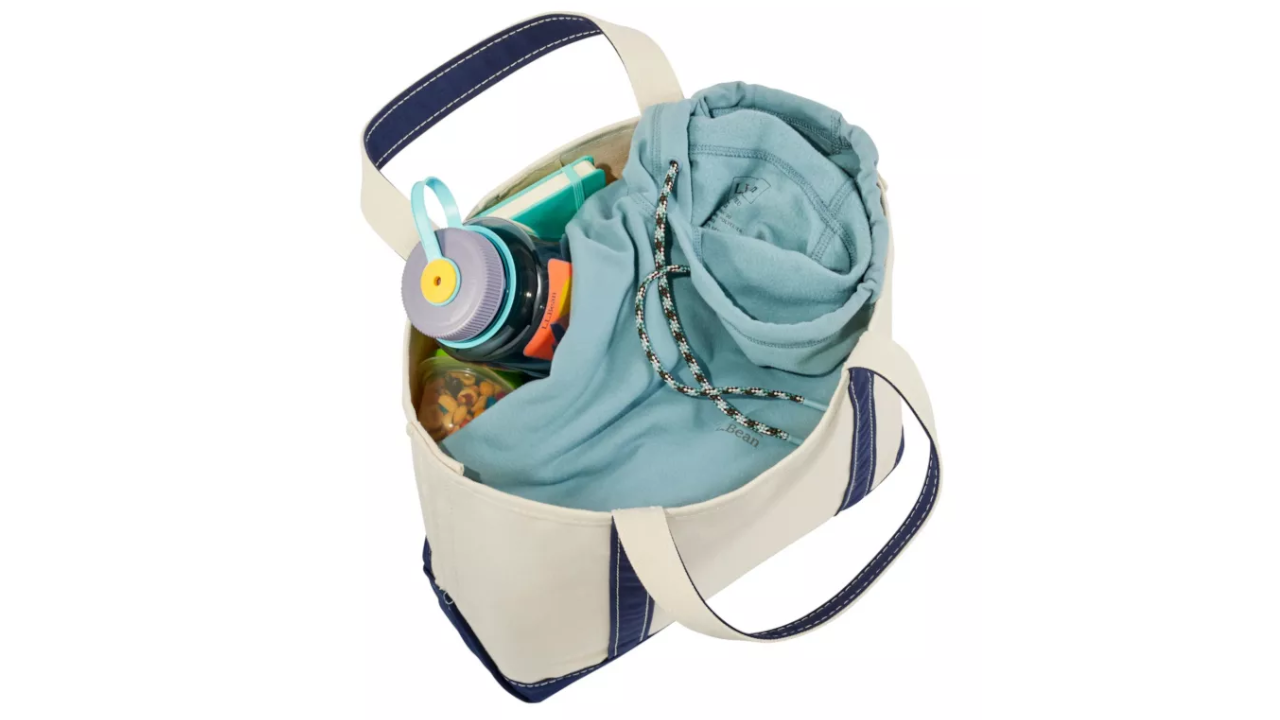 White canvas tote bag with navy blue trim containing a light blue hoodie, water bottle, and other personal items