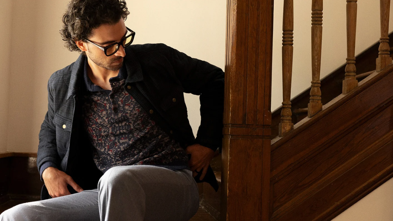 Man with curly hair and glasses wearing a patterned sweater and dark jacket sitting on wooden stairs