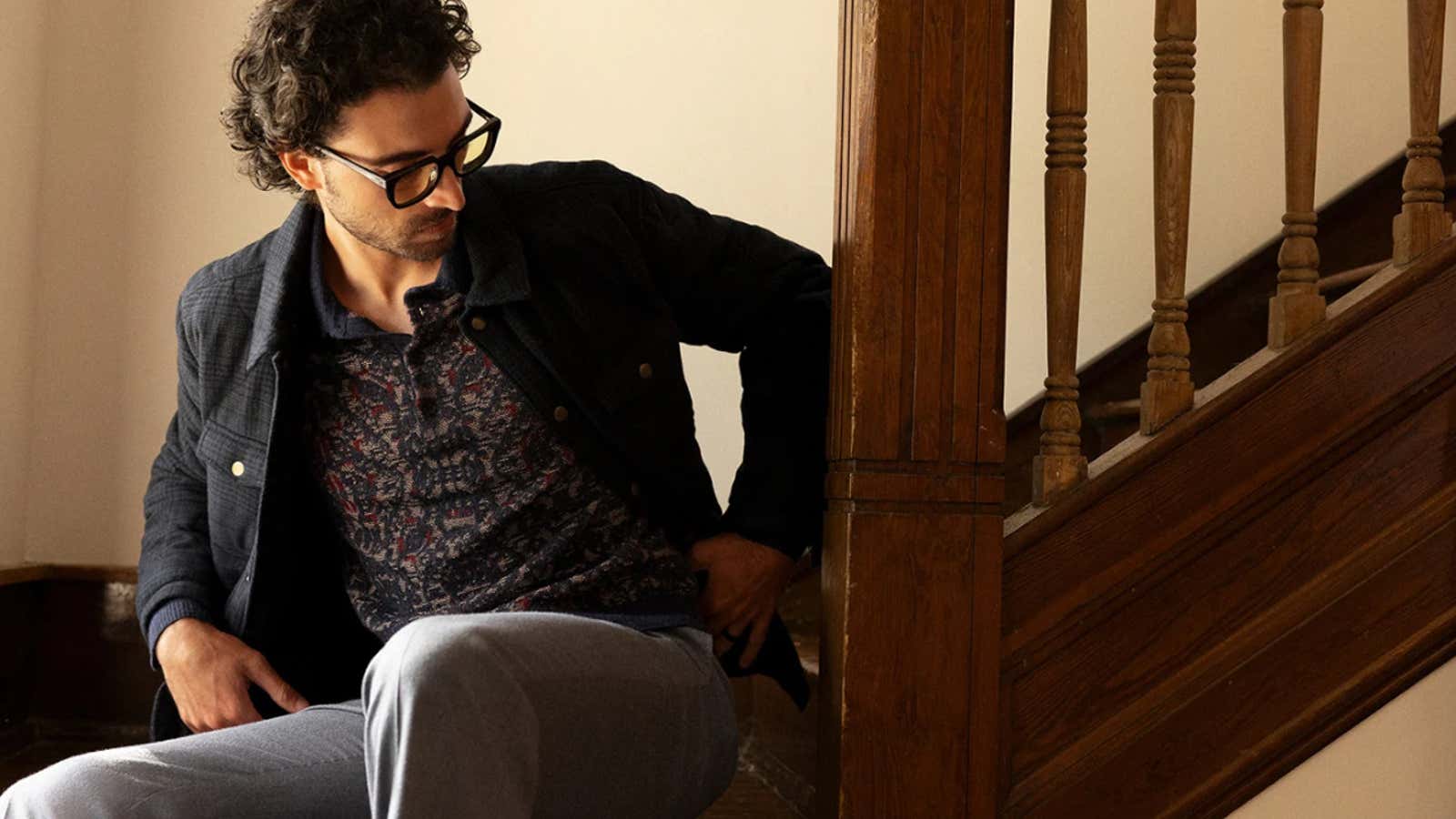 Man with curly hair and glasses wearing a patterned sweater and dark jacket sitting on wooden stairs