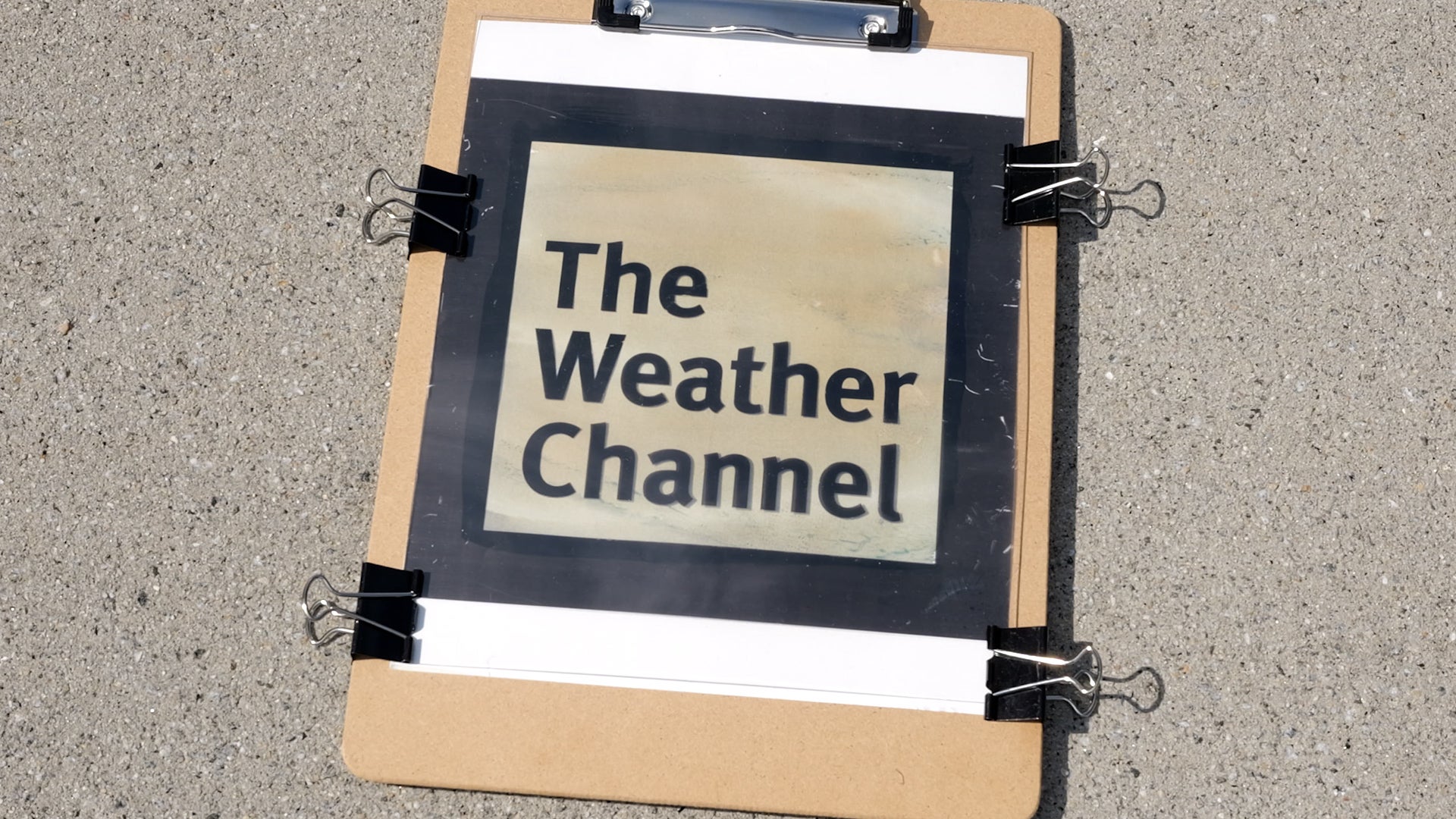A printed "The Weather Channel" logo attached to a clipboard with binder clips, resting on pavement