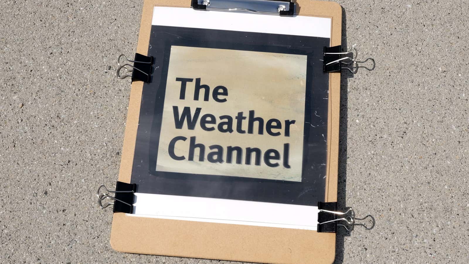 A printed "The Weather Channel" logo attached to a clipboard with binder clips, resting on pavement