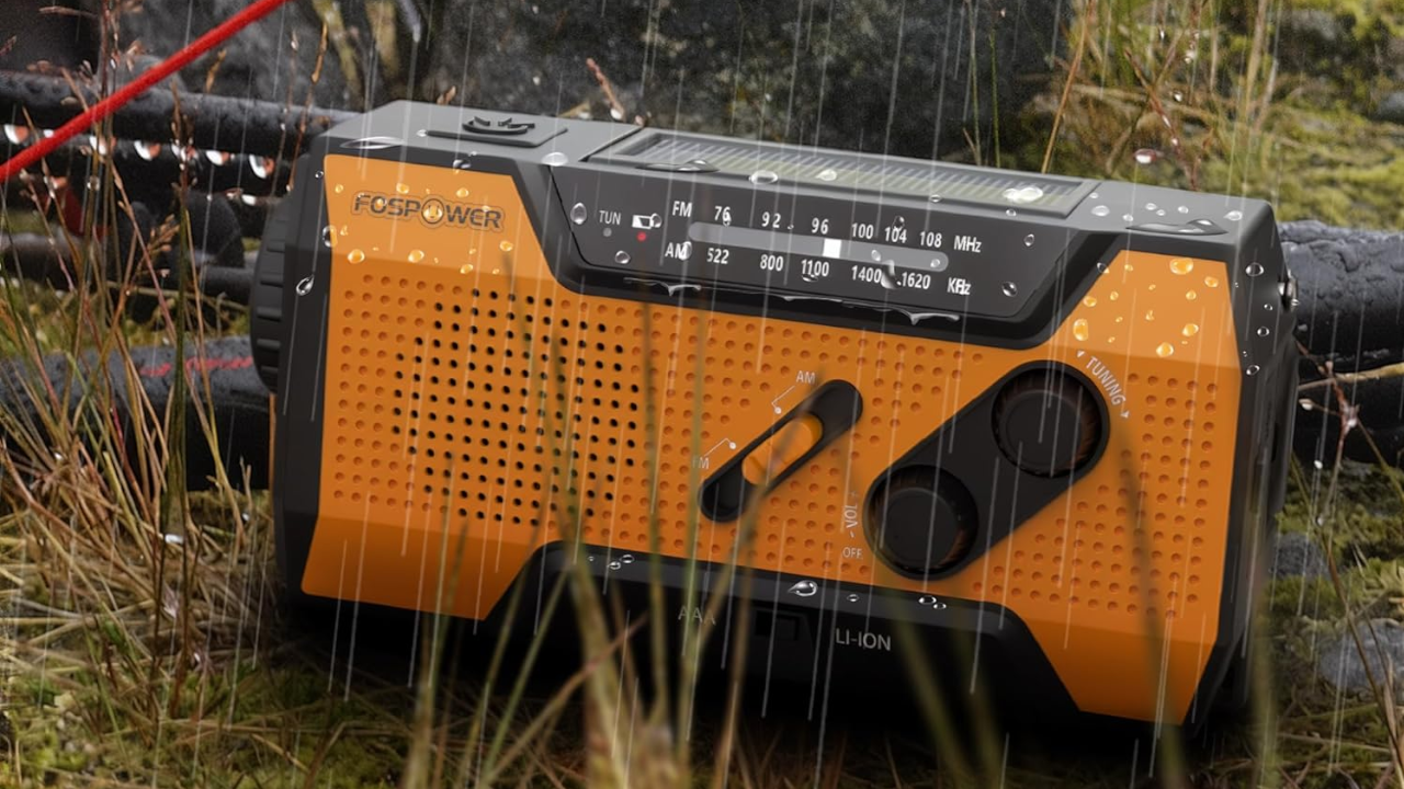 Orange and black FosPower emergency radio with AM/FM dial and hand crank sitting outdoors in tall grass during rainfall