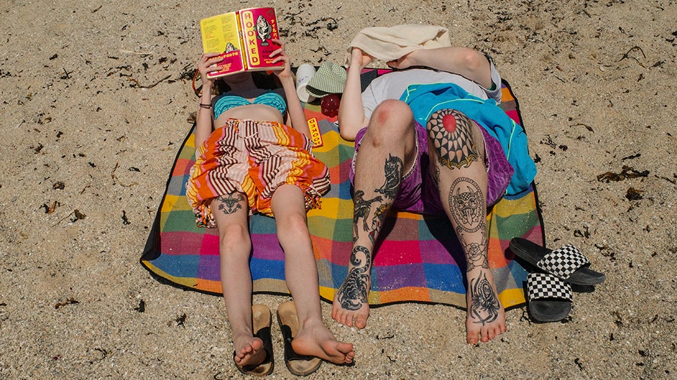 Two people lying on a beach, one reading a book.