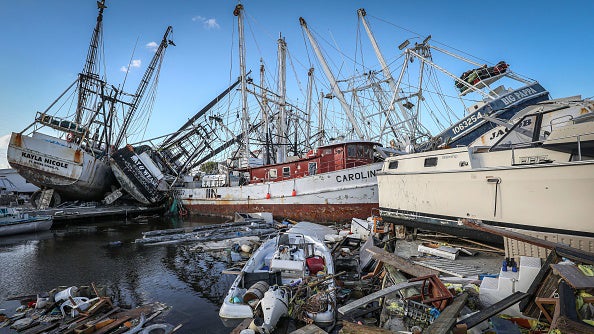 Damaged fishing boats and debris crowd a harbor after storm destruction, with vessels tilted and wreckage scattered in the water.