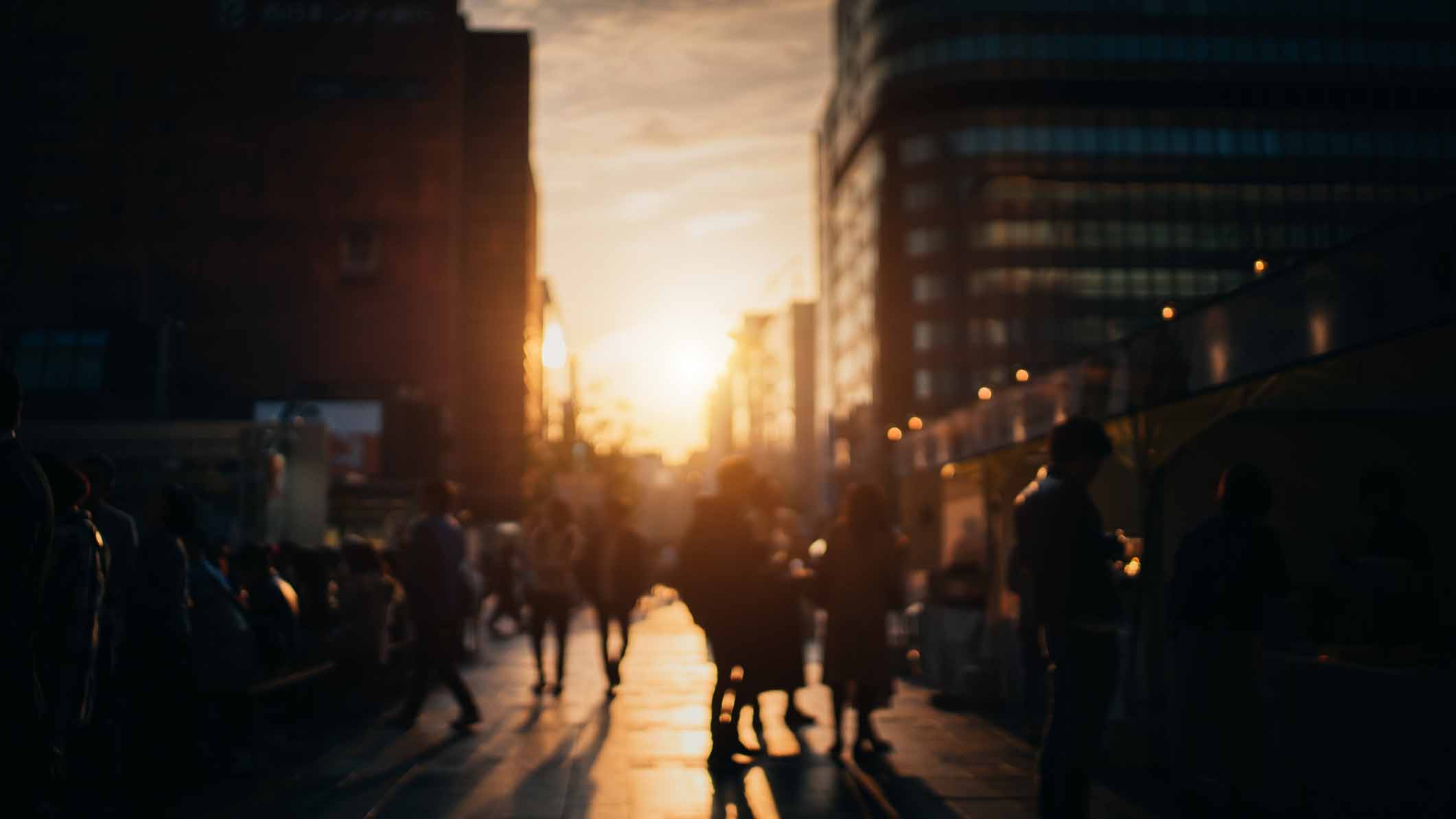 Blurred silhouettes of pedestrians walking on a city street as the sun sets between tall buildings.