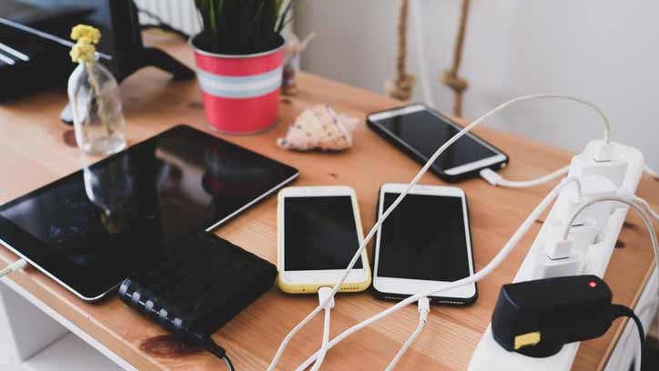Smartphones, a tablet, and a power bank charging on a wooden desk with a power strip, plant, and seashell.