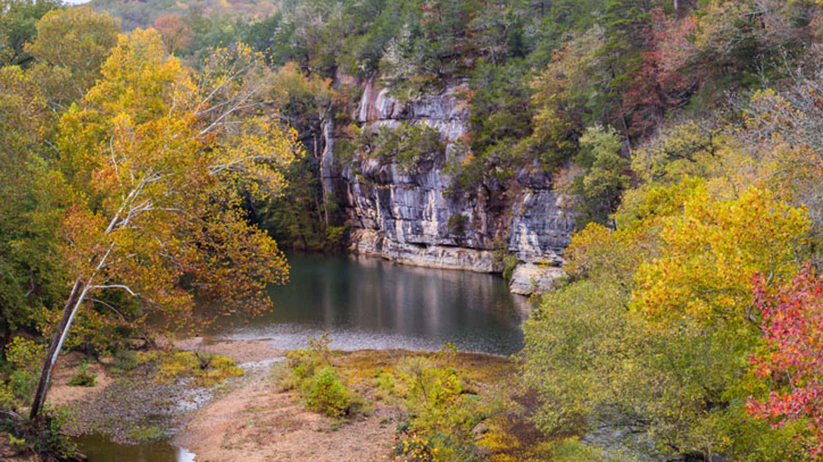 Rocky limestone bluff overlooking a calm river surrounded by colorful autumn foliage.