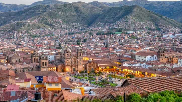 Aerial view of Cusco, Peru at dusk, showing the main plaza, cathedral, and hillside city surrounded by mountains.