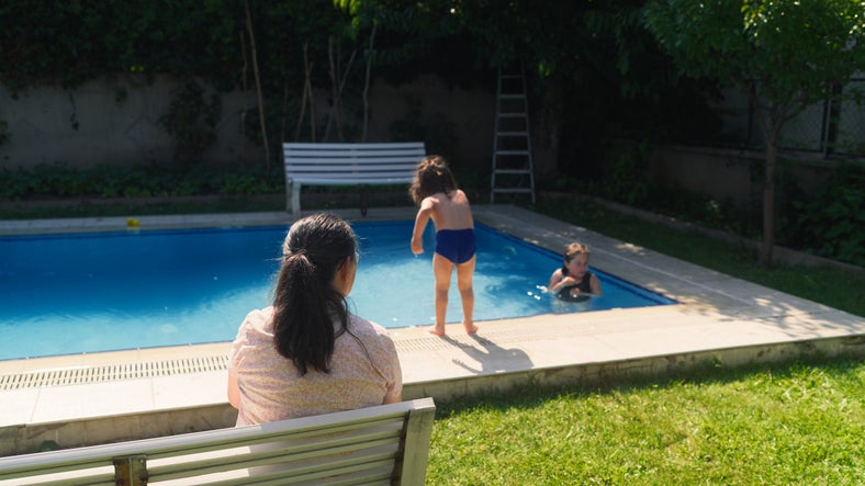 An adult woman sits by the pool where she's keeping a close eye on two children swimming.