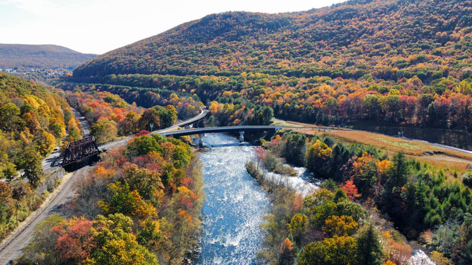 Aerial view of a river valley with a bridge, railroad tracks, and forested mountains showing vibrant autumn foliage.