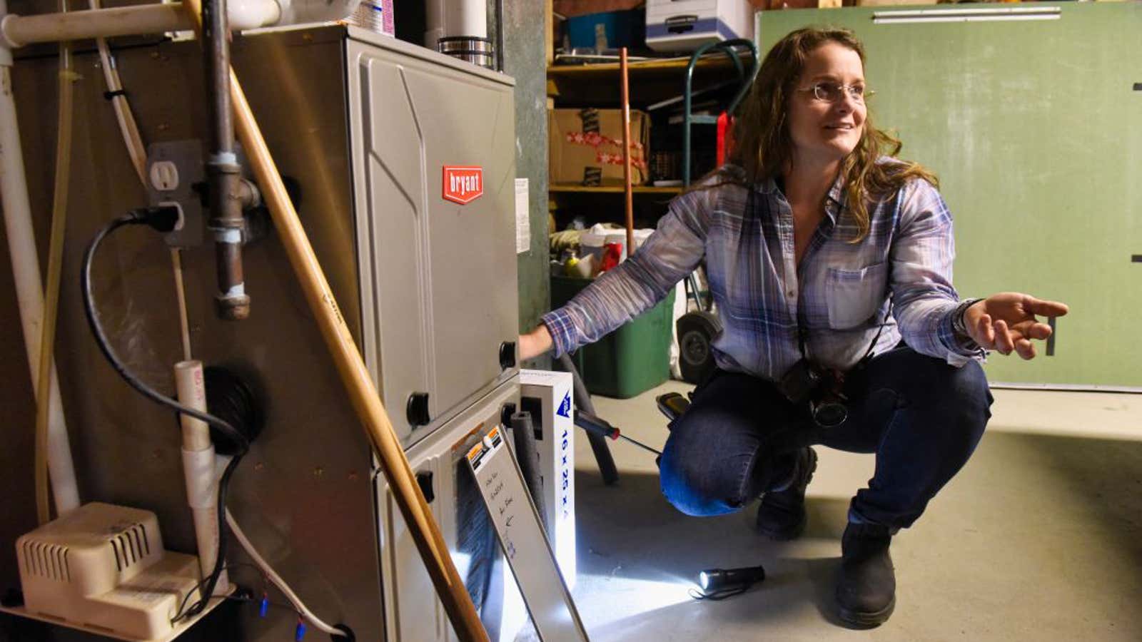 Woman in plaid shirt crouching beside a furnace, gesturing while inspecting the unit in a garage.