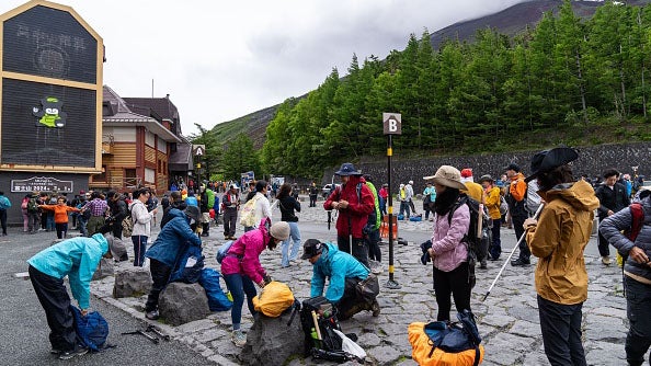 Hikers gather and prepare gear at a mountain trail base station, with a wooden building and green hillside in the background.