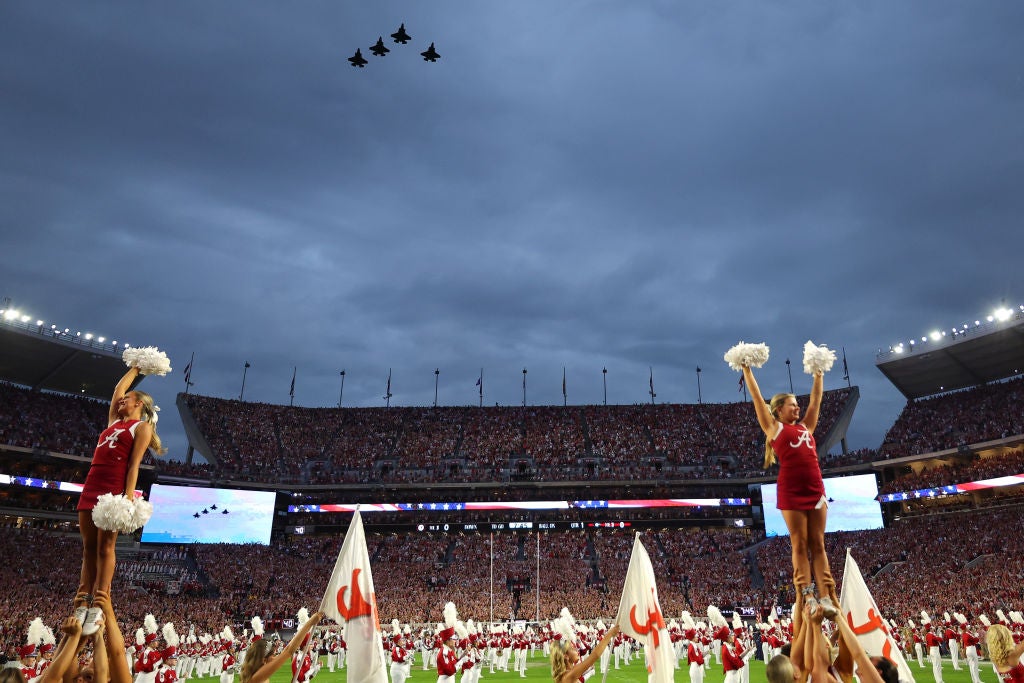 Military jets perform a flyover above a packed football stadium as cheerleaders are lifted during a pregame ceremony