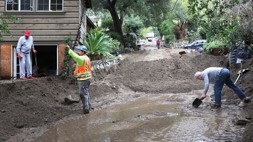 Residents shovel mud and debris from a flooded street after heavy rain, with damaged homes and cars nearby