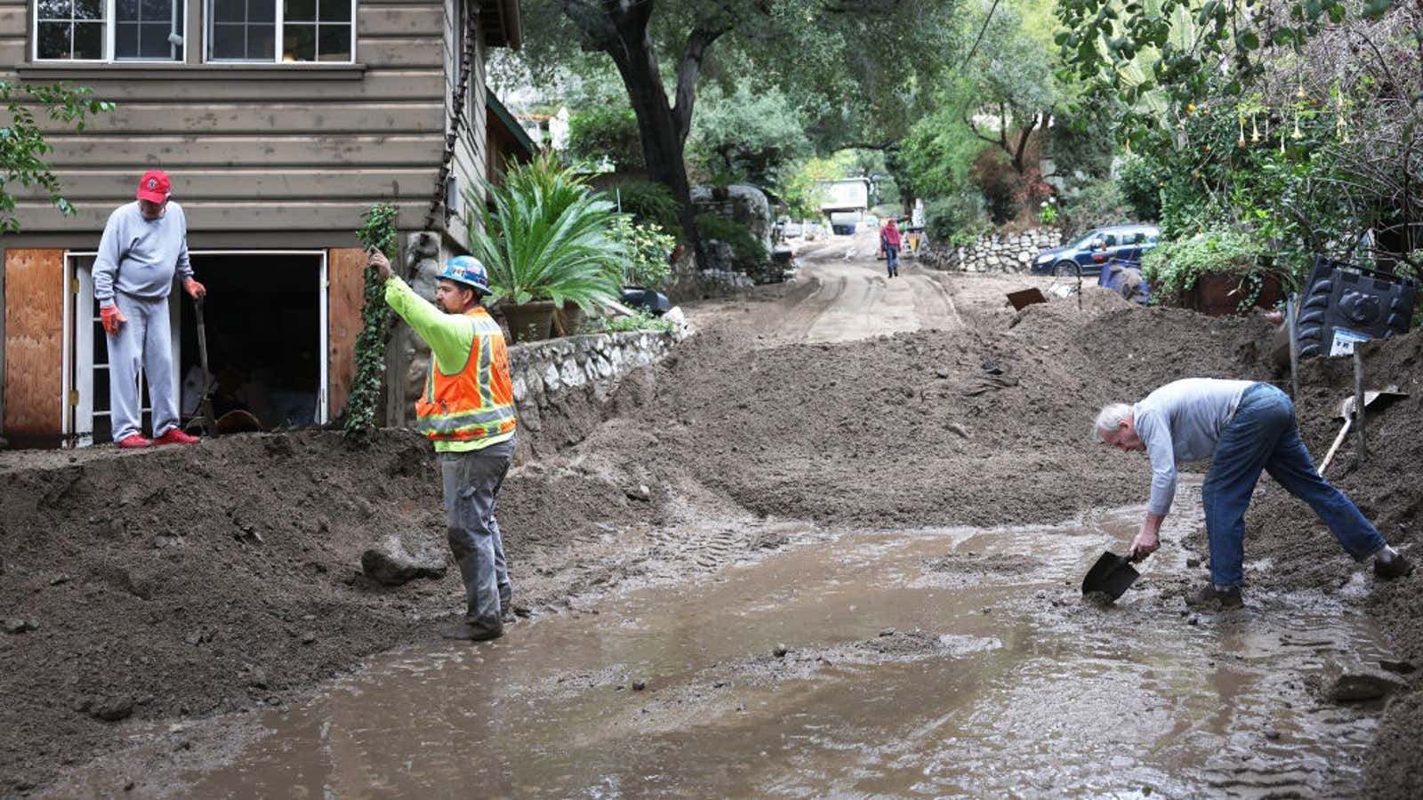 Residents shovel mud and debris from a flooded street after heavy rain, with damaged homes and cars nearby
