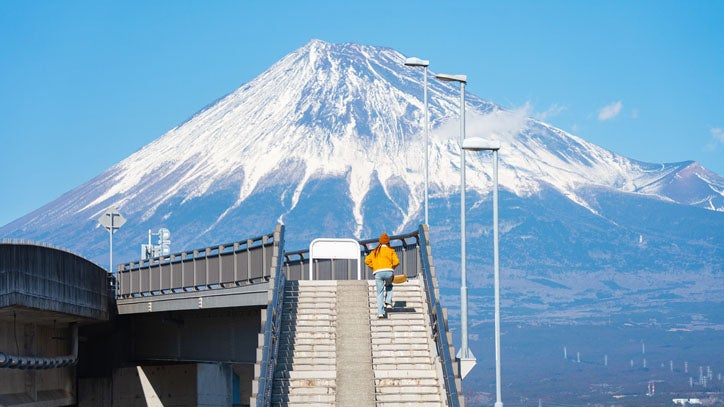 Person in orange jacket climbing steps on an overpass with snow-capped Mount Fuji in the background.
