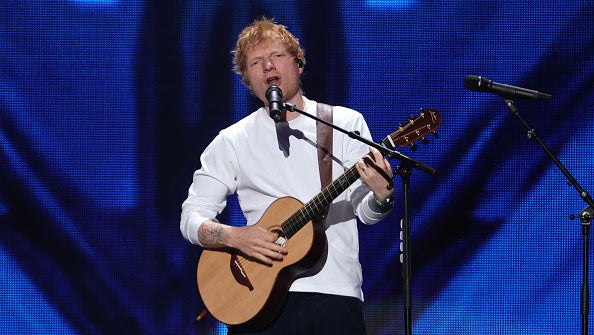 Male musician with red hair singing and playing acoustic guitar on stage with blue lighting background.