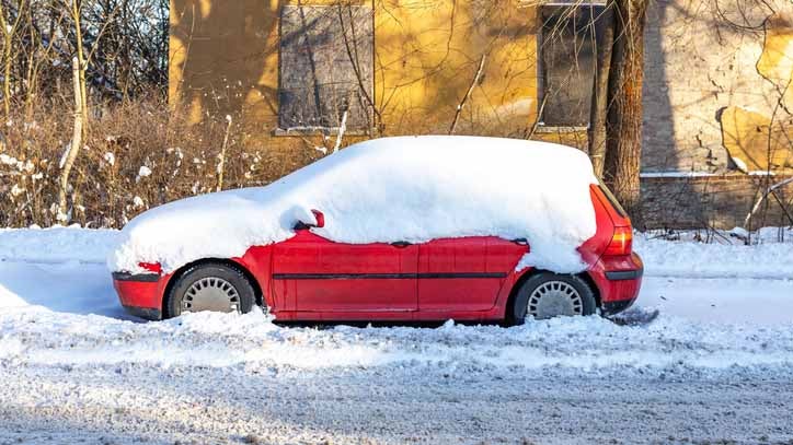 Red car covered in thick snow parked on a snowy street beside a yellow building.
