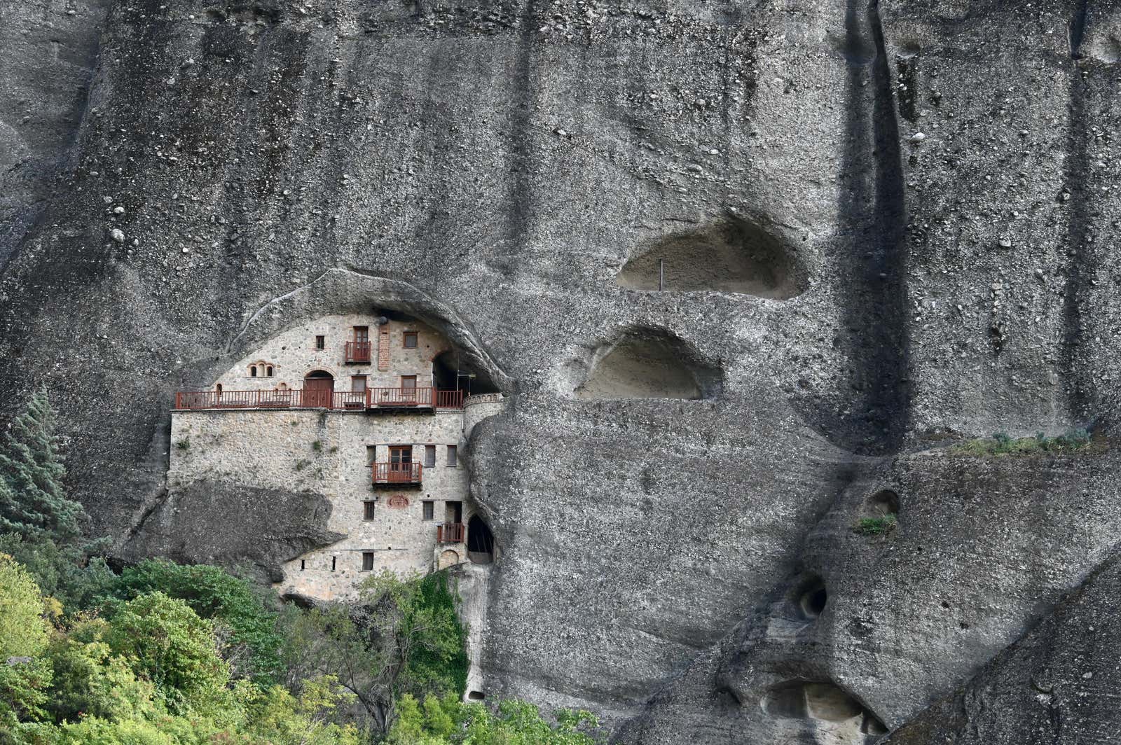 Stone monastery building built into a cave on a towering rocky cliff, surrounded by greenery at the base