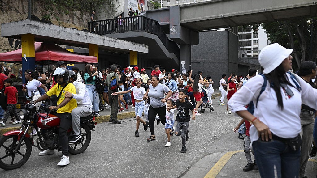 Large crowd of people running on a street under concrete overpass structures with motorcycles and urban buildings visible