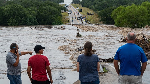 People stand on opposite sides of a road being washed out by an overflowing river.