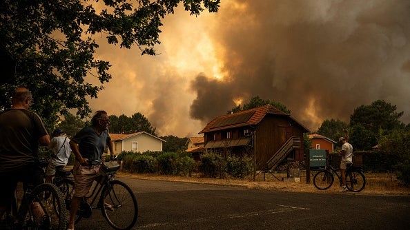 People with bicycles watch a huge smoke cloud rising behind houses in a neighborhood.