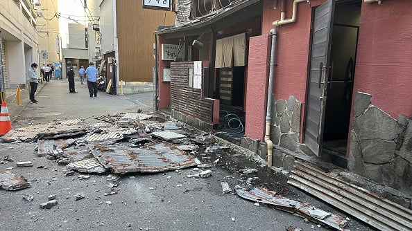Damaged building with debris and broken roofing scattered across a narrow street, people standing nearby.