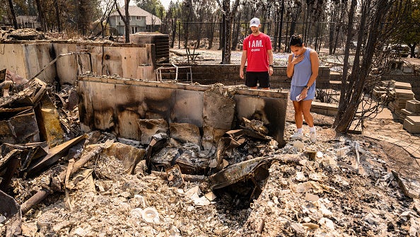 Two people stand amid the charred rubble of a house destroyed by wildfire, with burnt trees in the background.