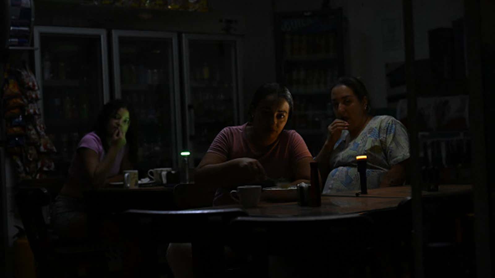 Three women sit at tables in a dimly lit shop illuminated by small green and orange indicator lights.